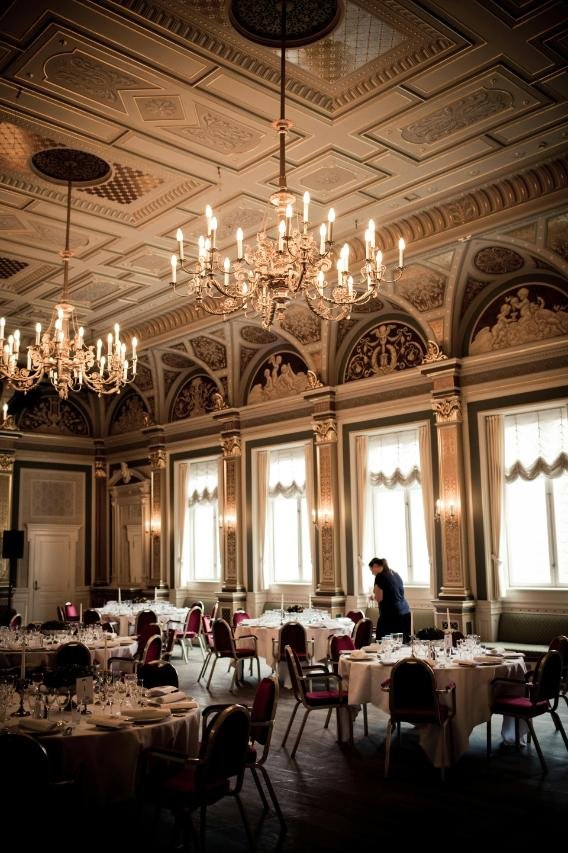 Elegant banquet hall with ornate ceiling, chandeliers, large windows, set tables, and a staff member preparing for an event.