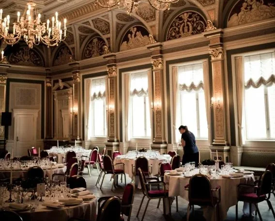 Elegant banquet hall with ornate ceilings, chandeliers, round tables, and a person preparing the setting. Perfect for events.