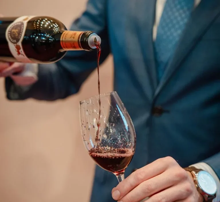 Man in a suit pouring red wine into a glass at a formal event; wine tasting or sommelier service concept.